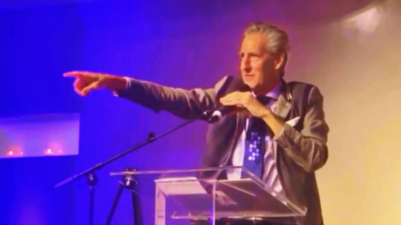Gene Seidman at a podium pointing into the audience during a charity auction, lit by blue and amber stage lights