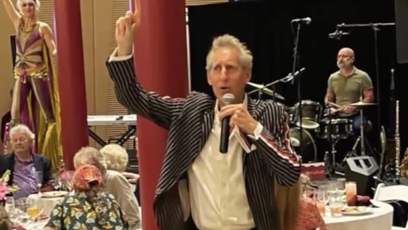 Gene Seidman working the crowd in a striped blazer, microphone in hand, arm raised at a lively charity event with a live band and performers in the background