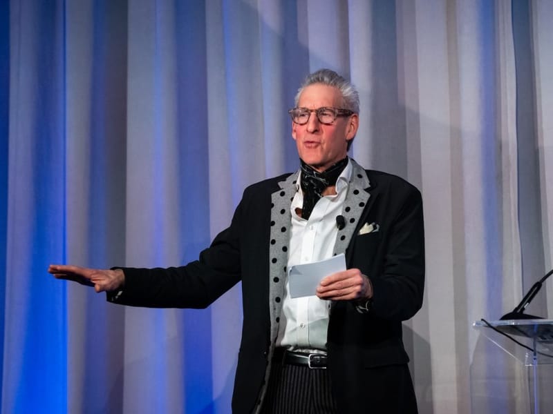 Gene Seidman on stage at a charity event, gesturing with one hand and holding note cards, wearing glasses and a patterned scarf