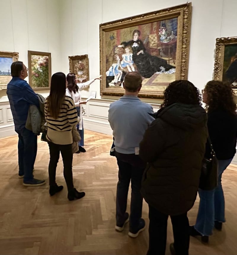 Museum guide pointing at a large Renoir portrait painting while a group of visitors listens in a gallery
