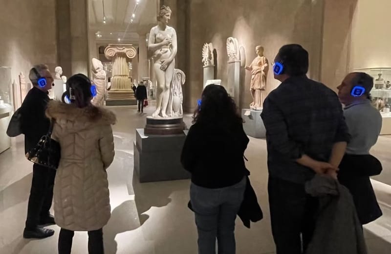 Group of museum visitors wearing blue wireless headsets studying a marble statue in a gallery of ancient Greek and Roman sculpture